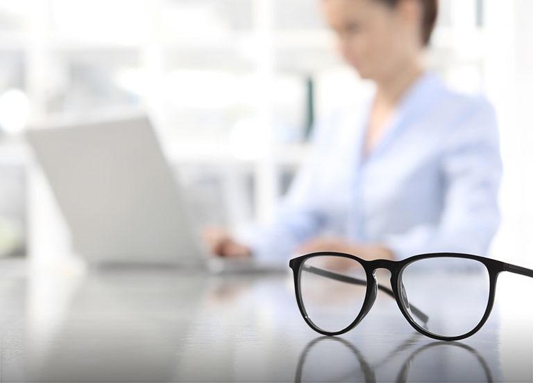 Okulary leżące na stole, kobieta pracująca przy laptopie
